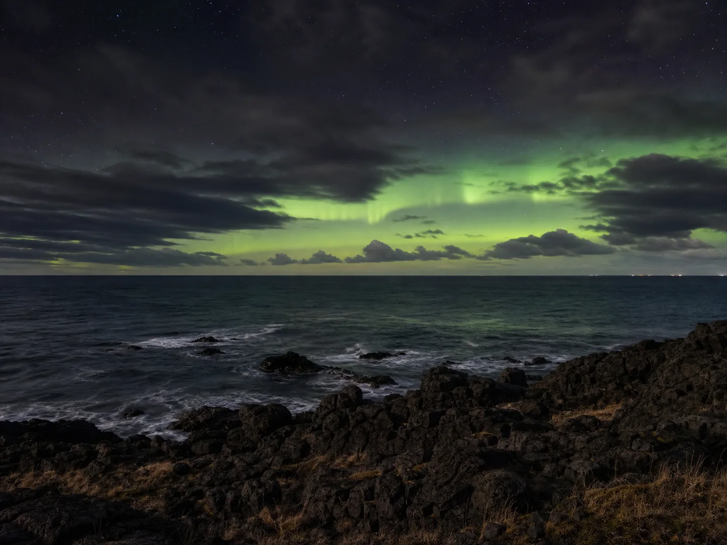 An AI-created green aurora above an open black-rock coast and low horizon